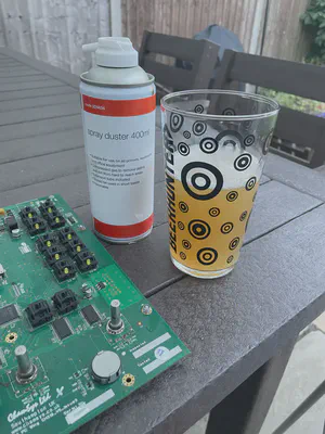 A circuit board from inside the PC Wing on a patio table, with a glass of beer and a can of spray cleaner